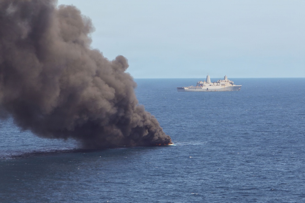 A vessel is actively burning on the open ocean, producing a large column of dark gray smoke rising into an overcast sky. In the background, a gray naval warship — appearing to be an amphibious transport dock or similar military vessel — is positioned at a distance observing or responding to the incident. Visible flames can be seen at the waterline of the burning vessel.