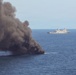 A vessel is actively burning on the open ocean, producing a large column of dark gray smoke rising into an overcast sky. In the background, a gray naval warship — appearing to be an amphibious transport dock or similar military vessel — is positioned at a distance observing or responding to the incident. Visible flames can be seen at the waterline of the burning vessel.