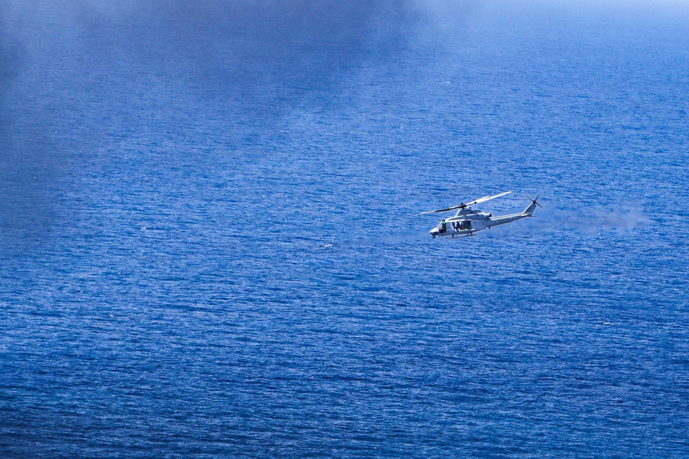 A military or coast guard helicopter is flying low over open ocean water. The helicopter appears to be a twin-rotor utility type, likely engaged in a patrol, search-and-rescue, or surveillance mission. The deep blue ocean surface dominates the frame, with the helicopter positioned in the center-right area of the image.