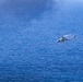 A military or coast guard helicopter is flying low over open ocean water. The helicopter appears to be a twin-rotor utility type, likely engaged in a patrol, search-and-rescue, or surveillance mission. The deep blue ocean surface dominates the frame, with the helicopter positioned in the center-right area of the image.