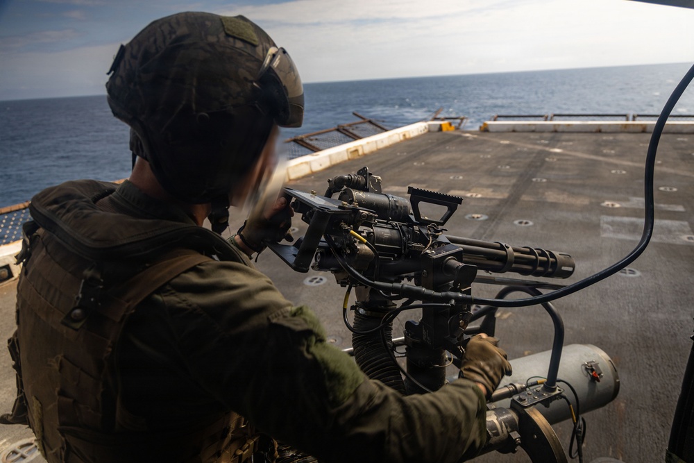 A military service member is positioned on what appears to be the deck of a naval vessel, operating a mounted multi-barrel rotary machine gun (consistent with an M134 Minigun or similar weapon system). The operator is wearing full combat gear including a helmet, tactical vest, and gloves, and is aiming the weapon toward the open ocean. The setting appears to be a ship's flight deck or open deck area during daylight hours.