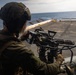 A military service member is positioned on what appears to be the deck of a naval vessel, operating a mounted multi-barrel rotary machine gun (consistent with an M134 Minigun or similar weapon system). The operator is wearing full combat gear including a helmet, tactical vest, and gloves, and is aiming the weapon toward the open ocean. The setting appears to be a ship's flight deck or open deck area during daylight hours.
