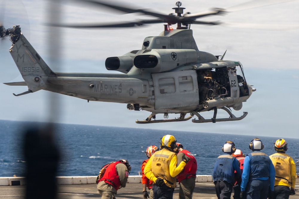 A U.S. Marines UH-1Y Venom helicopter (Bureau Number 168512) is hovering over the flight deck of a naval vessel, likely an amphibious assault ship. Deck crew members in color-coded safety gear are observing or directing the helicopter operation. The scene depicts a standard naval flight deck evolution over open ocean.
