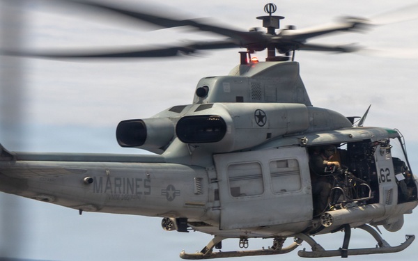 A U.S. Marines UH-1Y Venom helicopter (Bureau Number 168512) is hovering over the flight deck of a naval vessel, likely an amphibious assault ship. Deck crew members in color-coded safety gear are observing or directing the helicopter operation. The scene depicts a standard naval flight deck evolution over open ocean.