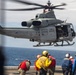 A U.S. Marines UH-1Y Venom helicopter (Bureau Number 168512) is hovering over the flight deck of a naval vessel, likely an amphibious assault ship. Deck crew members in color-coded safety gear are observing or directing the helicopter operation. The scene depicts a standard naval flight deck evolution over open ocean.