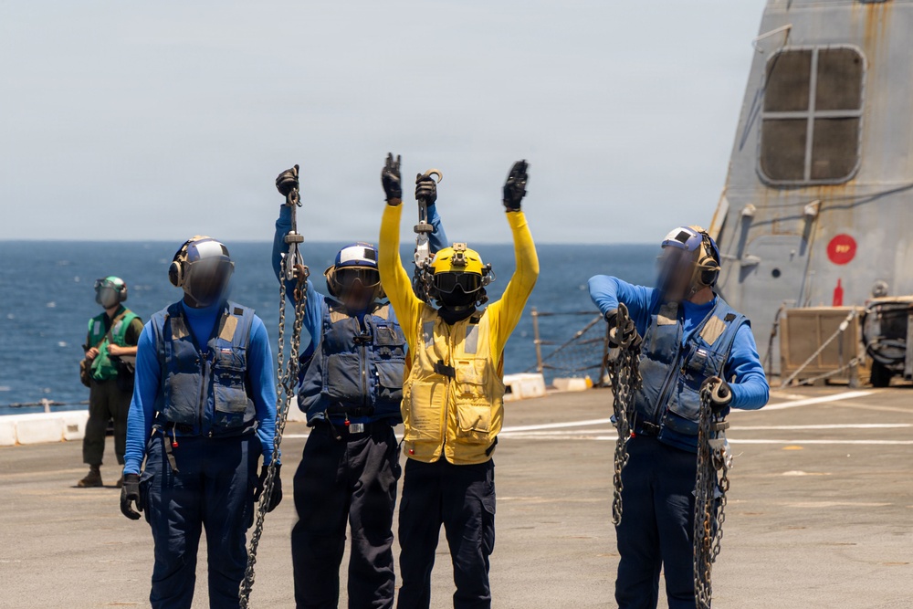 Naval personnel are conducting flight deck operations aboard what appears to be a U.S. Navy vessel at sea. Four crew members in protective gear are visible on the flight deck, with one individual in a yellow jersey raising both arms in a signaling gesture while others hold tie-down chains. A second ship is partially visible in the background to the right.