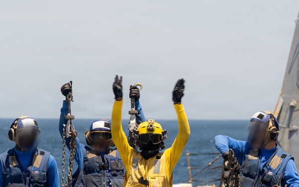 Naval personnel are conducting flight deck operations aboard what appears to be a U.S. Navy vessel at sea. Four crew members in protective gear are visible on the flight deck, with one individual in a yellow jersey raising both arms in a signaling gesture while others hold tie-down chains. A second ship is partially visible in the background to the right.