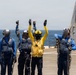 Naval personnel are conducting flight deck operations aboard what appears to be a U.S. Navy vessel at sea. Four crew members in protective gear are visible on the flight deck, with one individual in a yellow jersey raising both arms in a signaling gesture while others hold tie-down chains. A second ship is partially visible in the background to the right.