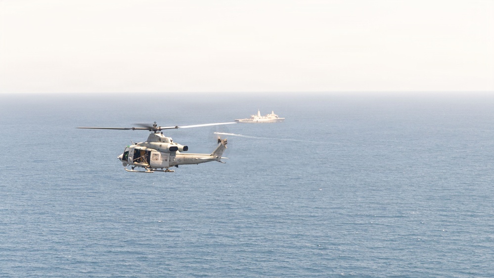 A military helicopter flies over open ocean with a naval vessel visible in the background. The helicopter appears to be a light utility or attack variant in a gray/white color scheme, consistent with U.S. Marine Corps or Navy markings. The scene suggests a maritime patrol or training operation.