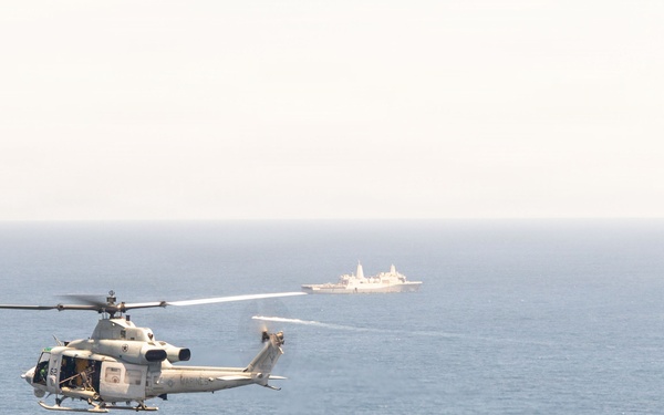 A military helicopter flies over open ocean with a naval vessel visible in the background. The helicopter appears to be a light utility or attack variant in a gray/white color scheme, consistent with U.S. Marine Corps or Navy markings. The scene suggests a maritime patrol or training operation.