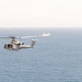 A military helicopter flies over open ocean with a naval vessel visible in the background. The helicopter appears to be a light utility or attack variant in a gray/white color scheme, consistent with U.S. Marine Corps or Navy markings. The scene suggests a maritime patrol or training operation.