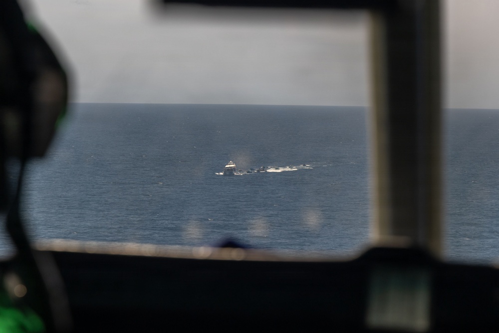 The image is taken from inside an aircraft cockpit looking out through the windshield at an open ocean scene. In the distance, at least one vessel is visible on the water, surrounded by what appears to be white water disturbance or wake. The foreground shows blurred cockpit framing and instrumentation.