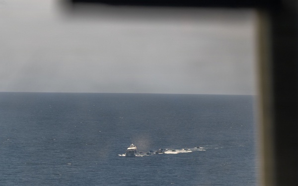 The image is taken from inside an aircraft cockpit looking out through the windshield at an open ocean scene. In the distance, at least one vessel is visible on the water, surrounded by what appears to be white water disturbance or wake. The foreground shows blurred cockpit framing and instrumentation.