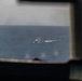 The image is taken from inside an aircraft cockpit looking out through the windshield at an open ocean scene. In the distance, at least one vessel is visible on the water, surrounded by what appears to be white water disturbance or wake. The foreground shows blurred cockpit framing and instrumentation.