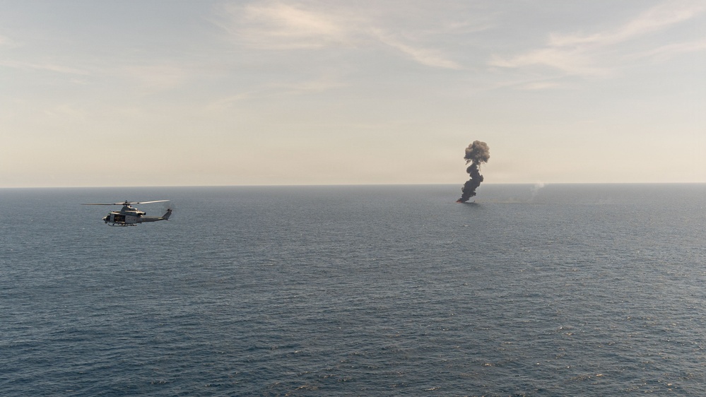 A military helicopter flies over open ocean near a large column of dark black smoke rising from the water's surface. The smoke appears to originate from a burning vessel or target at sea. The scene is consistent with a military strike, weapons test, or controlled destruction exercise.