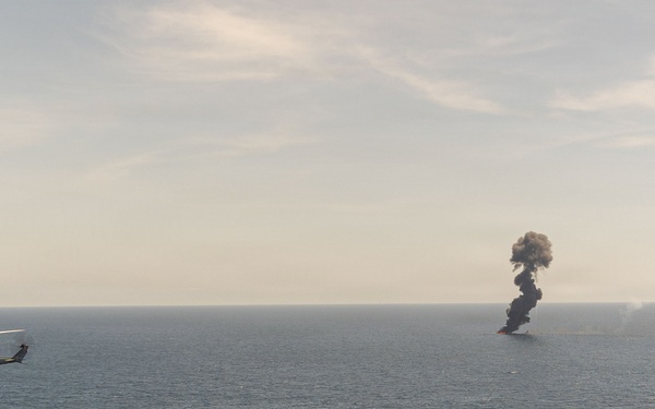 A military helicopter flies over open ocean near a large column of dark black smoke rising from the water's surface. The smoke appears to originate from a burning vessel or target at sea. The scene is consistent with a military strike, weapons test, or controlled destruction exercise.