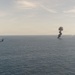 A military helicopter flies over open ocean near a large column of dark black smoke rising from the water's surface. The smoke appears to originate from a burning vessel or target at sea. The scene is consistent with a military strike, weapons test, or controlled destruction exercise.
