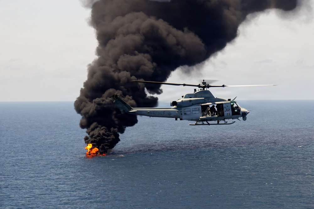 A U.S. Marines military helicopter, marked with the number '62' and 'MARINES' on its fuselage, hovers over open ocean near a burning fire on the water's surface. Large plumes of thick black smoke rise dramatically from the flames below. This appears to be a military exercise or interdiction operation over open sea.