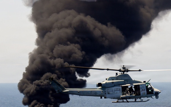A U.S. Marines military helicopter, marked with the number '62' and 'MARINES' on its fuselage, hovers over open ocean near a burning fire on the water's surface. Large plumes of thick black smoke rise dramatically from the flames below. This appears to be a military exercise or interdiction operation over open sea.