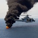A U.S. Marines military helicopter, marked with the number '62' and 'MARINES' on its fuselage, hovers over open ocean near a burning fire on the water's surface. Large plumes of thick black smoke rise dramatically from the flames below. This appears to be a military exercise or interdiction operation over open sea.