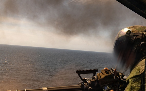 A military aircrew member is operating a multi-barrel rotary cannon (consistent with an M134 Minigun) from the open door of a helicopter over a body of water. The weapon appears to be actively firing, evidenced by motion blur and smoke visible near the muzzle area. Large plumes of dark and white smoke are visible on the horizon in the background.