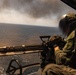 A military aircrew member is operating a multi-barrel rotary cannon (consistent with an M134 Minigun) from the open door of a helicopter over a body of water. The weapon appears to be actively firing, evidenced by motion blur and smoke visible near the muzzle area. Large plumes of dark and white smoke are visible on the horizon in the background.