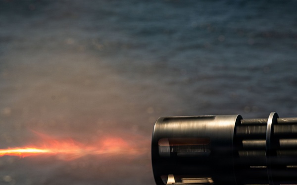 The image depicts a close-up of a gun barrel firing, with a visible muzzle flash and tracer round or projectile streak extending to the left. Smoke and combustion gases are visible around the barrel. The background shows a body of water, suggesting a maritime or naval setting.