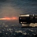 The image depicts a close-up of a gun barrel firing, with a visible muzzle flash and tracer round or projectile streak extending to the left. Smoke and combustion gases are visible around the barrel. The background shows a body of water, suggesting a maritime or naval setting.