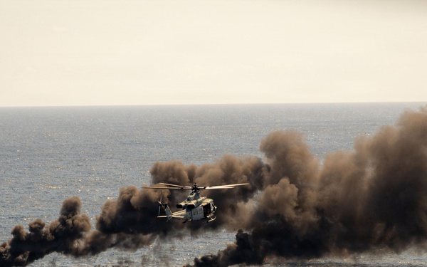 A military helicopter flies over open ocean near multiple small burning vessels or targets producing large columns of black and brown smoke. The scene appears to depict a maritime strike or interdiction exercise, with visible flames and heavy smoke rising from at least three surface targets. The image captures a dynamic military operation over open water under hazy sky conditions.