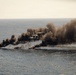 A military helicopter flies over open ocean near multiple small burning vessels or targets producing large columns of black and brown smoke. The scene appears to depict a maritime strike or interdiction exercise, with visible flames and heavy smoke rising from at least three surface targets. The image captures a dynamic military operation over open water under hazy sky conditions.