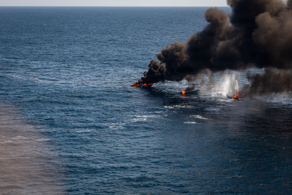 An aerial photograph shows multiple small vessels on fire in open ocean waters. Large plumes of dark black smoke rise from at least three burning boats aligned in a row. A white smoke or steam column is also visible on the rightmost vessel, suggesting an active fire suppression attempt or explosive event.