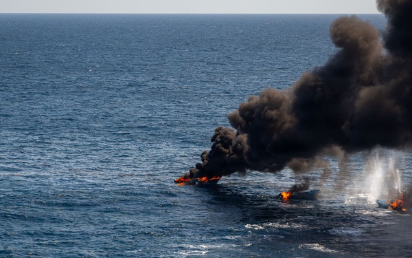 An aerial photograph shows multiple small vessels on fire in open ocean waters. Large plumes of dark black smoke rise from at least three burning boats aligned in a row. A white smoke or steam column is also visible on the rightmost vessel, suggesting an active fire suppression attempt or explosive event.