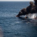 An aerial photograph shows multiple small vessels on fire in open ocean waters. Large plumes of dark black smoke rise from at least three burning boats aligned in a row. A white smoke or steam column is also visible on the rightmost vessel, suggesting an active fire suppression attempt or explosive event.
