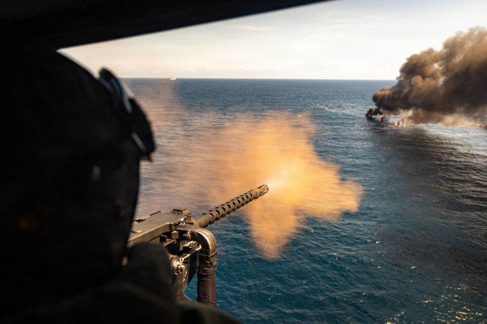 An aerial gunner is firing a mounted machine gun from what appears to be a military helicopter over open ocean. In the distance, a vessel or target is visibly burning and producing a large column of dark smoke. The perspective suggests this is taken from inside the aircraft during an active firing exercise or engagement.
