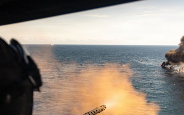 An aerial gunner is firing a mounted machine gun from what appears to be a military helicopter over open ocean. In the distance, a vessel or target is visibly burning and producing a large column of dark smoke. The perspective suggests this is taken from inside the aircraft during an active firing exercise or engagement.