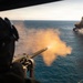 An aerial gunner is firing a mounted machine gun from what appears to be a military helicopter over open ocean. In the distance, a vessel or target is visibly burning and producing a large column of dark smoke. The perspective suggests this is taken from inside the aircraft during an active firing exercise or engagement.