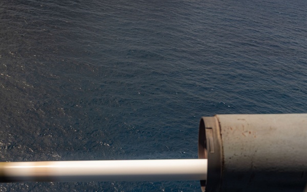 A large-caliber naval gun is captured mid-fire from an elevated shipboard position over open ocean. A projectile is visibly in motion exiting the muzzle, captured by high-speed photography with a bright muzzle flash/tracer effect. The image appears to be taken during a live-fire exercise at sea.