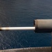 A large-caliber naval gun is captured mid-fire from an elevated shipboard position over open ocean. A projectile is visibly in motion exiting the muzzle, captured by high-speed photography with a bright muzzle flash/tracer effect. The image appears to be taken during a live-fire exercise at sea.
