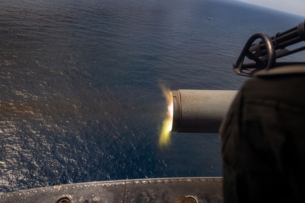 The image shows a large-caliber gun barrel firing over an open ocean, captured from an elevated aerial or shipboard perspective. A muzzle flash is clearly visible at the barrel's end, indicating active discharge. A small vessel or object is faintly visible on the water's surface in the distant background.