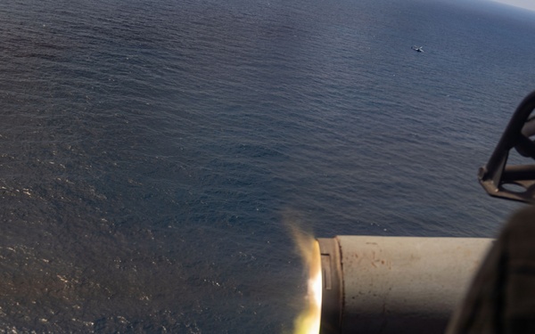 The image shows a large-caliber gun barrel firing over an open ocean, captured from an elevated aerial or shipboard perspective. A muzzle flash is clearly visible at the barrel's end, indicating active discharge. A small vessel or object is faintly visible on the water's surface in the distant background.