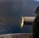 The image shows a large-caliber gun barrel firing over an open ocean, captured from an elevated aerial or shipboard perspective. A muzzle flash is clearly visible at the barrel's end, indicating active discharge. A small vessel or object is faintly visible on the water's surface in the distant background.