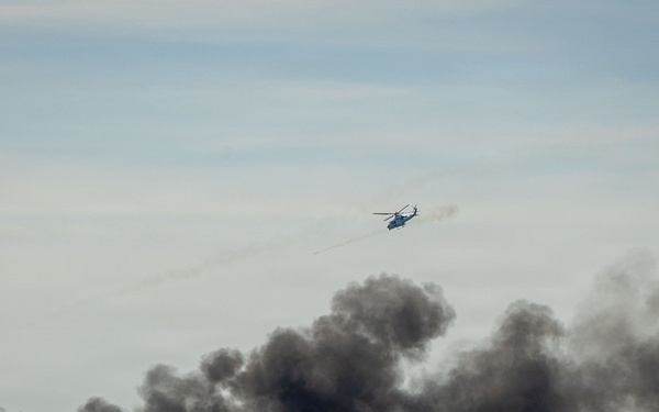 A military or emergency response helicopter is flying over an open body of water while large plumes of dark black smoke rise from what appears to be burning tires or debris on the water's surface. The helicopter appears to be deploying flares or conducting a firing exercise. This appears to be a military exercise or emergency response drill conducted at sea.