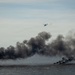 A military or emergency response helicopter is flying over an open body of water while large plumes of dark black smoke rise from what appears to be burning tires or debris on the water's surface. The helicopter appears to be deploying flares or conducting a firing exercise. This appears to be a military exercise or emergency response drill conducted at sea.