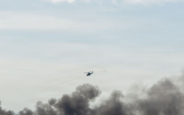 A military or emergency scenario over a body of water showing multiple sources of dark smoke rising from the water surface. A helicopter is visible in the sky above the smoke clouds, appearing to be conducting an operation or observation. The scene suggests either a naval exercise, combat engagement, or maritime incident involving burning vessels or targets.