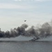 A military or emergency scenario over a body of water showing multiple sources of dark smoke rising from the water surface. A helicopter is visible in the sky above the smoke clouds, appearing to be conducting an operation or observation. The scene suggests either a naval exercise, combat engagement, or maritime incident involving burning vessels or targets.