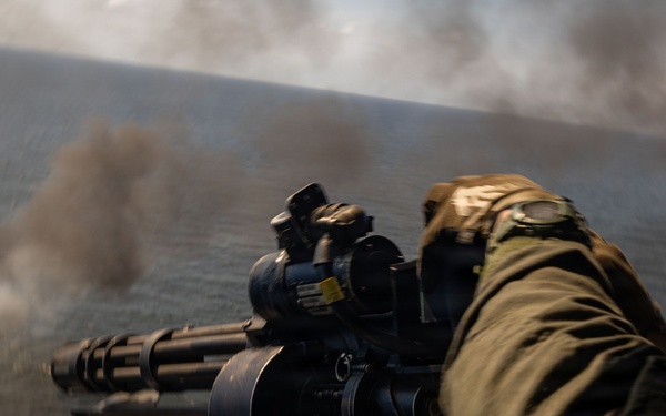 A military personnel in olive/green flight suit is operating a multi-barrel rotary cannon (consistent with an M134 Minigun) from what appears to be a helicopter or aerial platform over a body of water. Smoke or muzzle discharge is visible in the background, suggesting the weapon is being or has recently been fired. The perspective indicates this is an aerial gunnery operation.