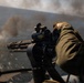 A military personnel in olive/green flight suit is operating a multi-barrel rotary cannon (consistent with an M134 Minigun) from what appears to be a helicopter or aerial platform over a body of water. Smoke or muzzle discharge is visible in the background, suggesting the weapon is being or has recently been fired. The perspective indicates this is an aerial gunnery operation.