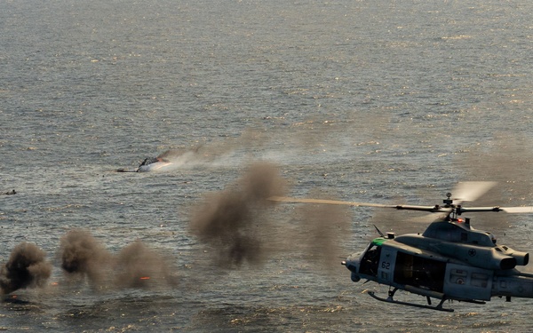 A military helicopter, appearing to be a UH-1Y Venom or similar twin-engine utility helicopter, is flying low over open water while engaging a surface target. Multiple explosion plumes and smoke clouds are visible on the water surface to the left of the aircraft, indicating an active weapons engagement or strike exercise. The scene appears to be a live-fire training exercise or combat operation over open ocean.