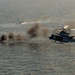 A military helicopter, appearing to be a UH-1Y Venom or similar twin-engine utility helicopter, is flying low over open water while engaging a surface target. Multiple explosion plumes and smoke clouds are visible on the water surface to the left of the aircraft, indicating an active weapons engagement or strike exercise. The scene appears to be a live-fire training exercise or combat operation over open ocean.