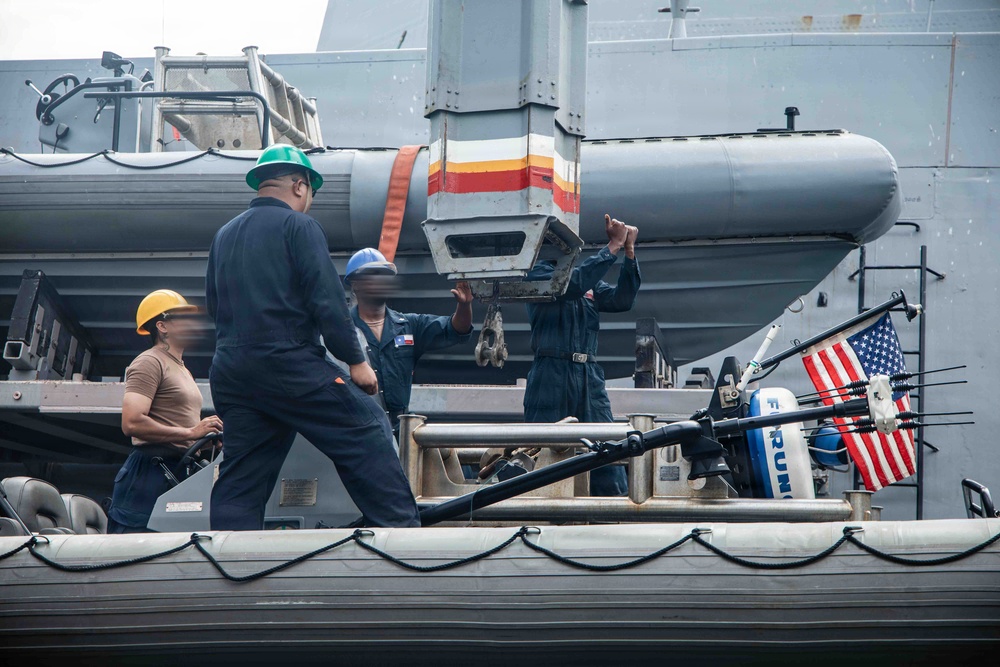 The image shows U.S. Navy personnel conducting deck operations aboard a naval vessel, likely during a boat handling or equipment maintenance evolution. Several sailors wearing different colored hard hats are working together on what appears to be a rigid hull inflatable boat (RHIB) or similar small craft secured to the ship's side. An American flag is visible in the background, confirming the U.S. military context.